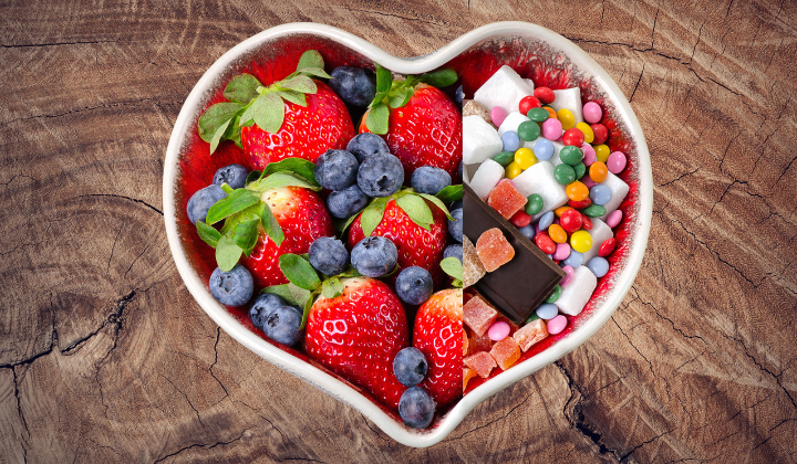 Umgang mit Fruktosemalabsorption Weisse Herzschale. Die eine Haelfte ist gefuellt mit Erdbeeren, Blaubeeren und die andere Haelfte mit Suessigkeiten. Die Schale steht auf einem Holztisch. Alles aus Vogelperspektive.